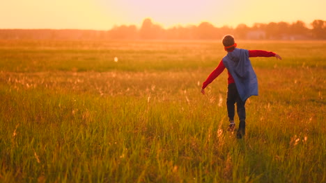 A-boy-in-a-suit-and-a-superhero-mask-with-a-red-cloak-runs-across-the-field-at-sunset-on-the-grass-dreaming-and-imagining-himself-a-hero-and-a-winner.